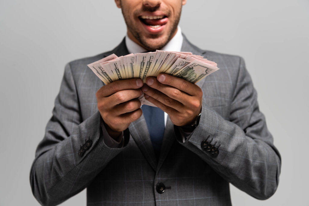 cropped view of businessman in suit holding dollar banknotes and licking lips isolated on grey